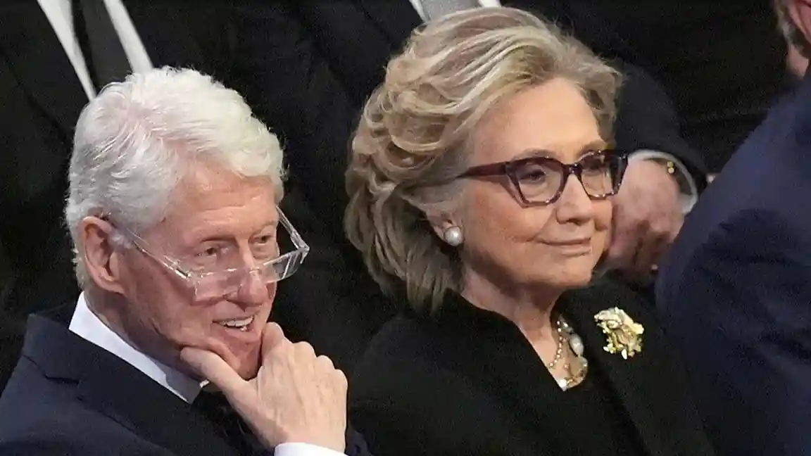 Bill and Hillary Clinton seated together at an event, with Bill Clinton smiling and resting his chin on his hand, and Hillary Clinton with glasses and a brooch, looking ahead.