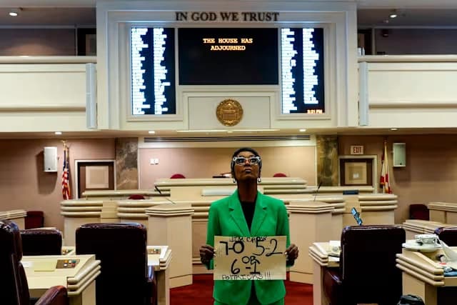 Woman holding a protest sign inside a legislative chamber during the Alabama redistricting debate.