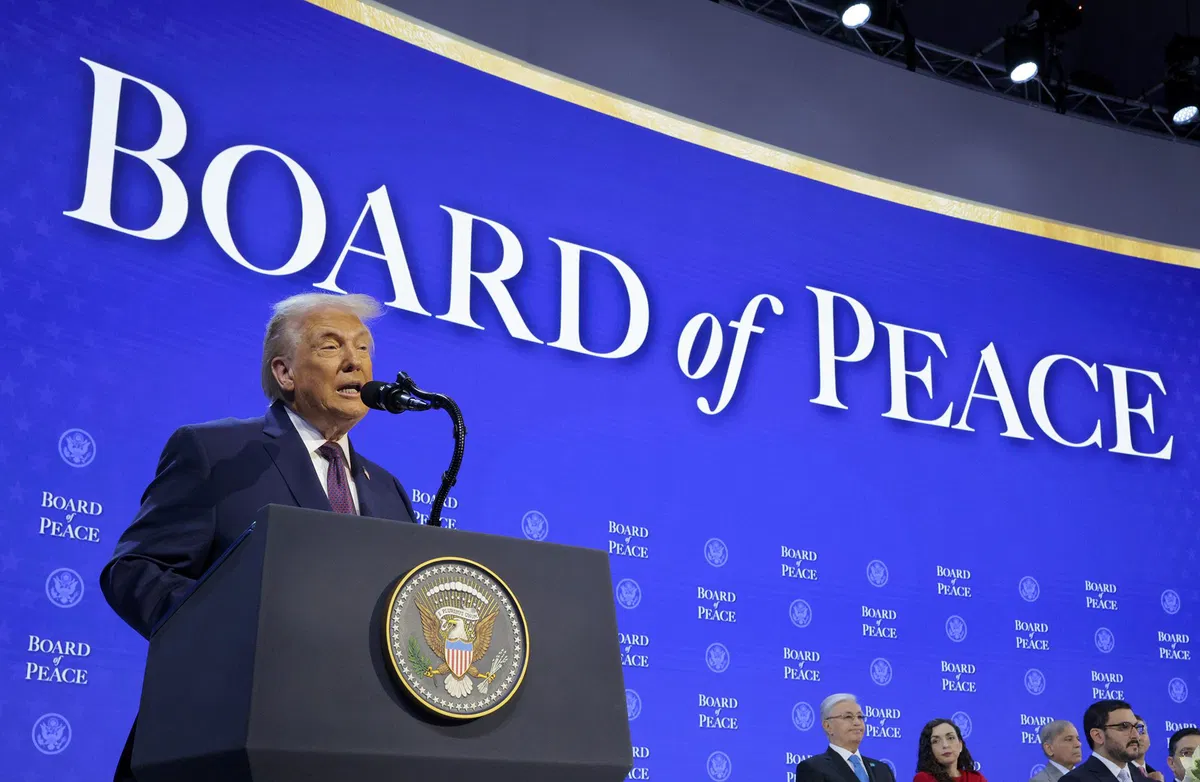 President Donald Trump speaks at a podium during the inaugural Board of Peace meeting, with international delegates seated behind him.